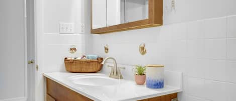 A bathroom with a white sink, wooden vanity, and a mirror. There is a basket on the sink, a potted plant, and a decorative container.