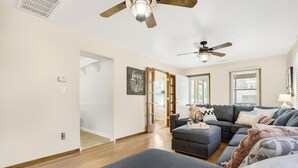 A spacious living room with a grey sectional sofa, a wooden floor, ceiling fans, and a large window with blinds.