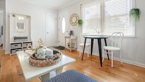 A bright, white-walled room with a wooden floor, a round table with two white chairs, a bench, and a small side table with a basket.