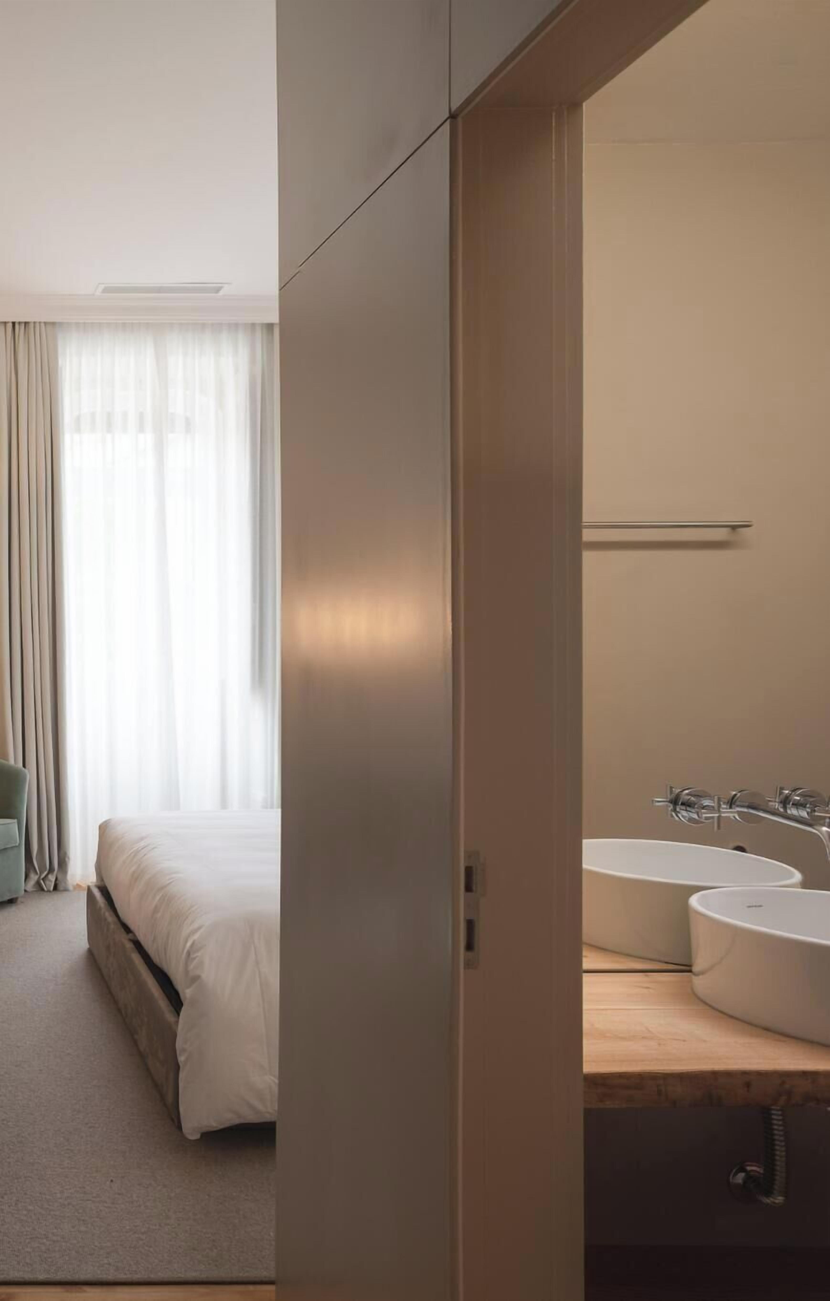 A hotel room with a bed, a large window with curtains, and a bathroom with a sink and mirror.