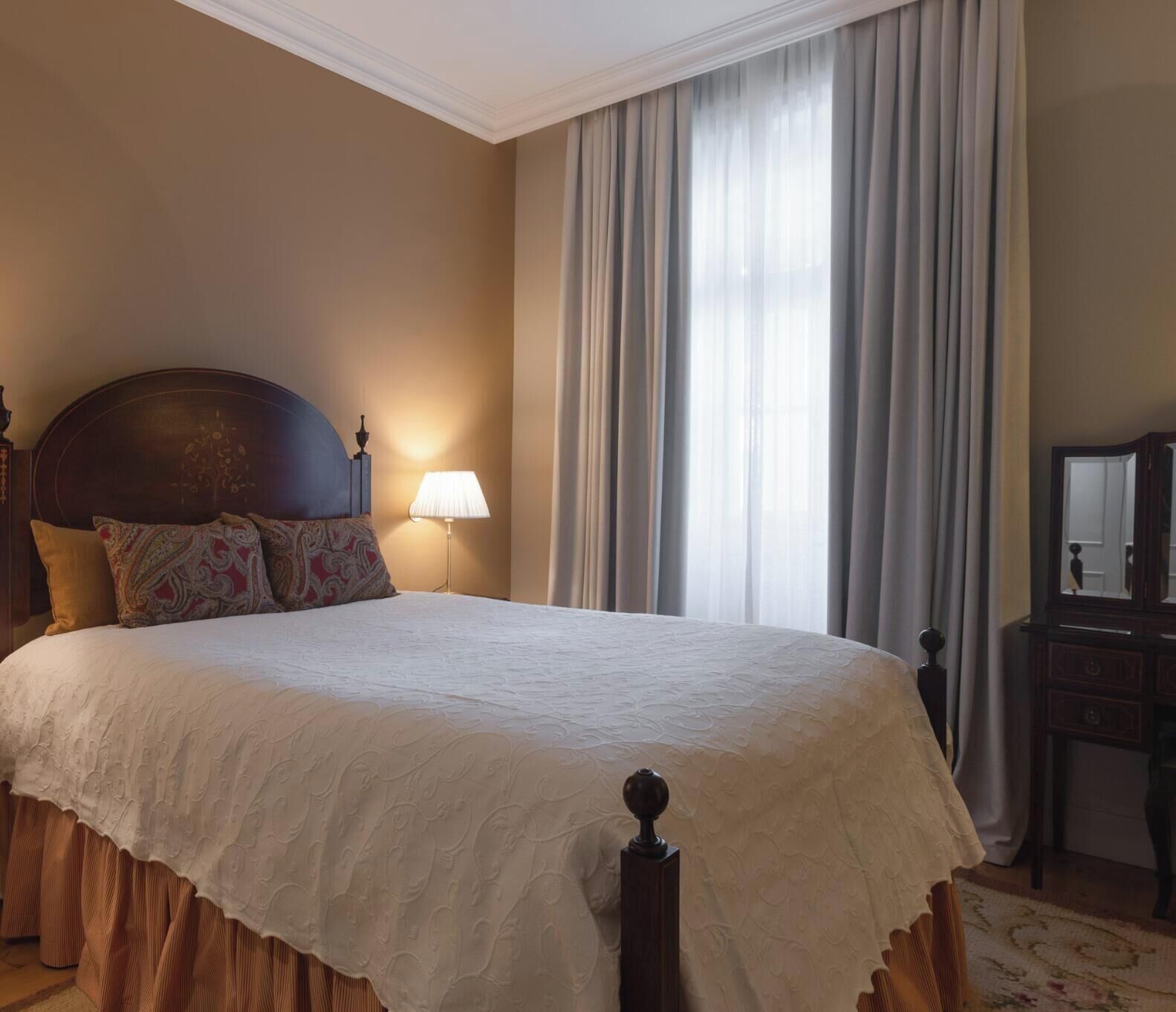 Un lit bien fait, avec une couette blanche, une tête de lit en bois et des coussins à motifs. On y trouve une lampe de chevet et une table de chevet en bois avec un miroir.