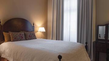 Un lit bien fait, avec une couette blanche, une tête de lit en bois et des coussins à motifs. On y trouve une lampe de chevet et une table de chevet en bois avec un miroir.