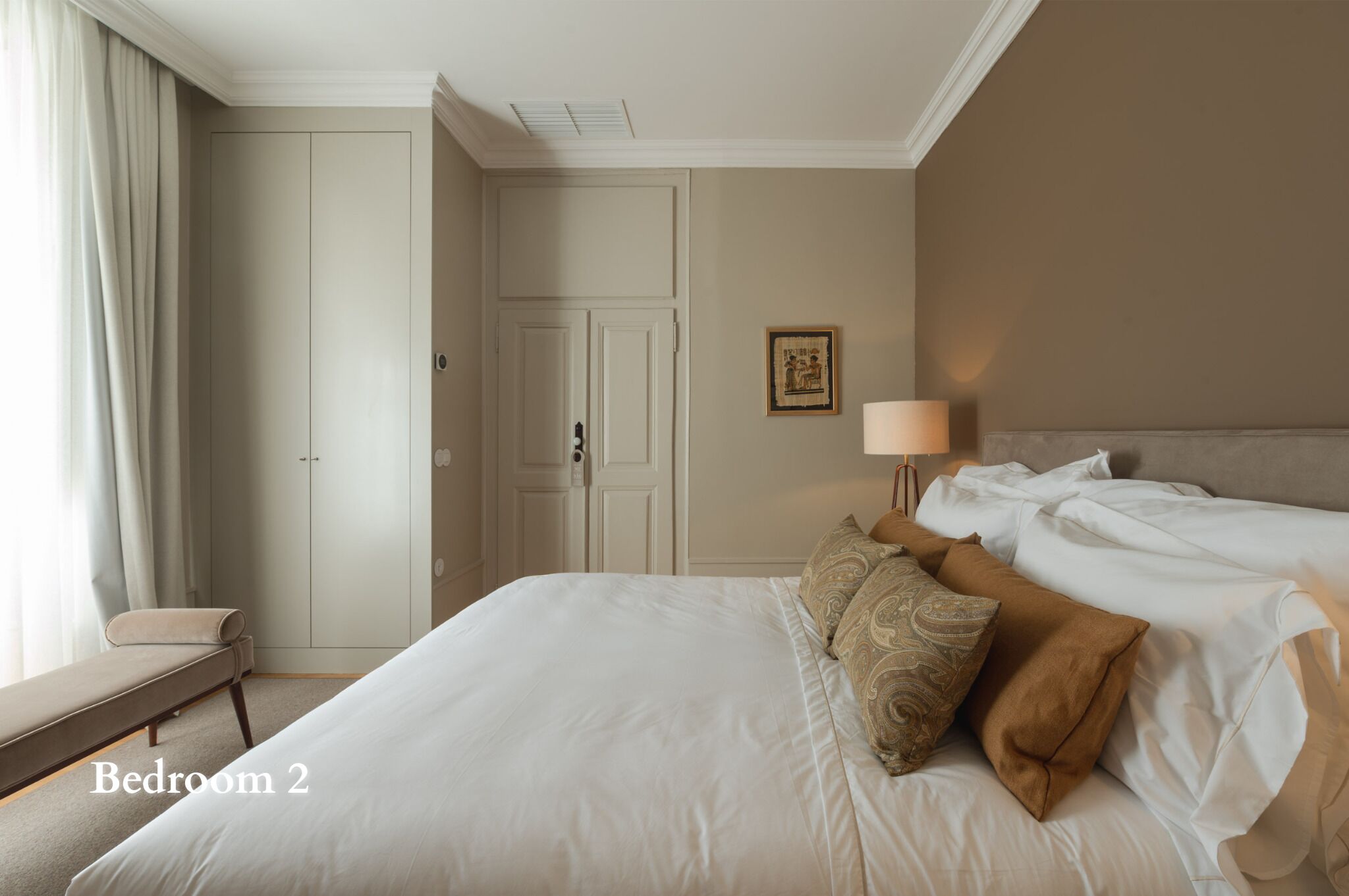 A bedroom with a bed, pillows, a nightstand, a lamp, a chair, and a door.