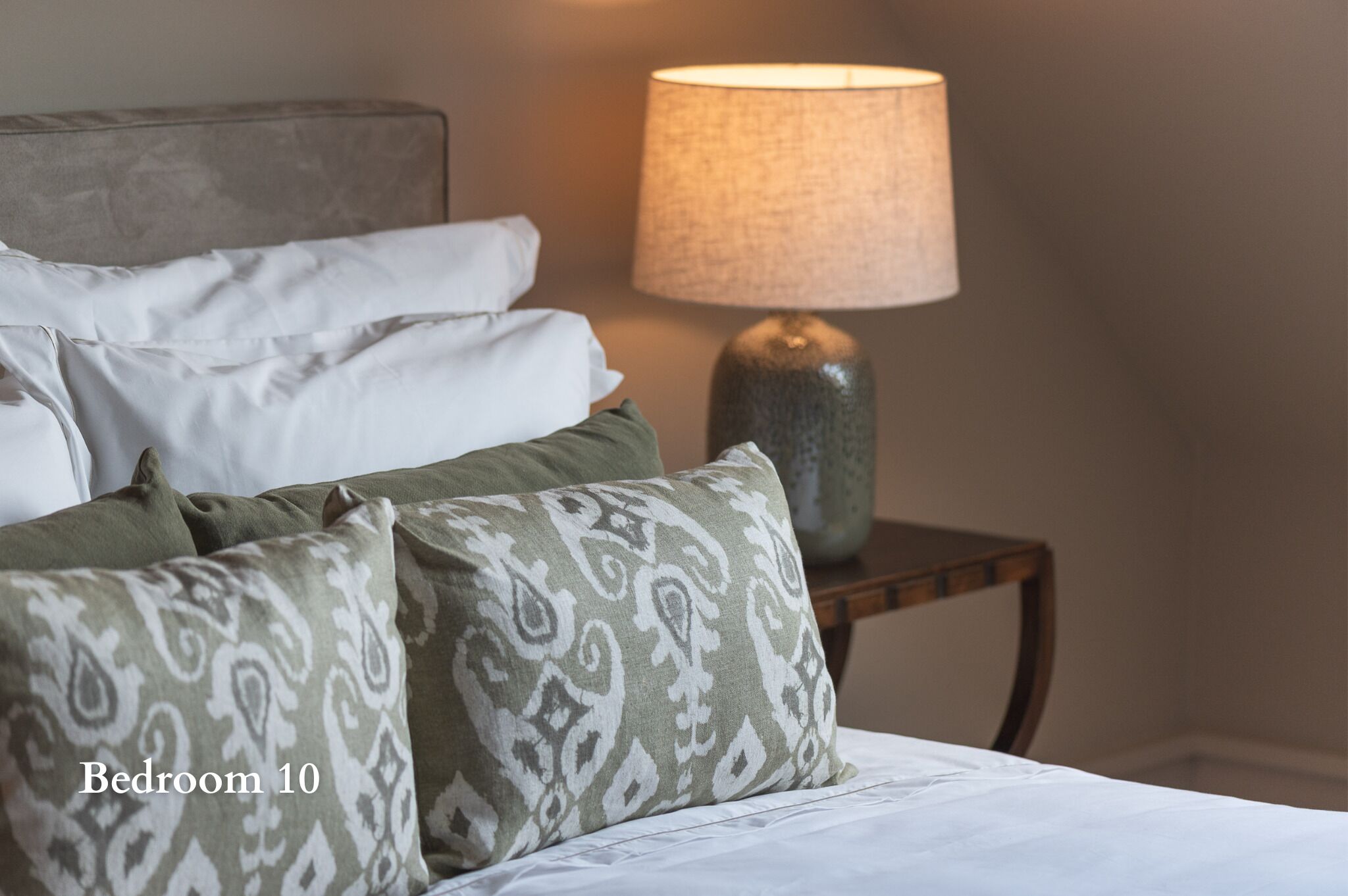 Un dormitorio con cama, almohadas, lámpara y mesita de noche.