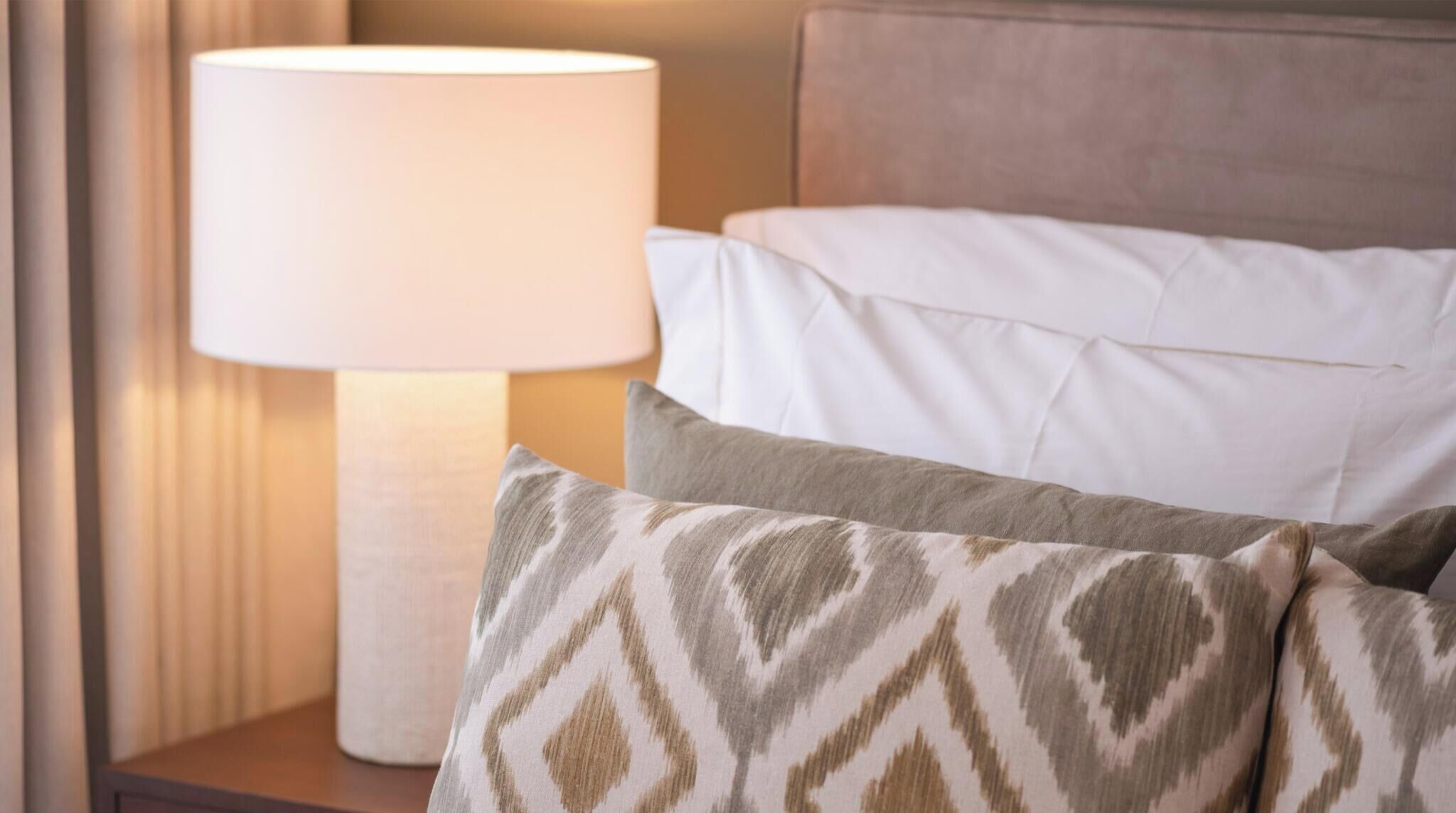 Een net opgemaakt bed met wit beddengoed en een kussen met een patroon, een bedlampje en een houten nachtkastje.