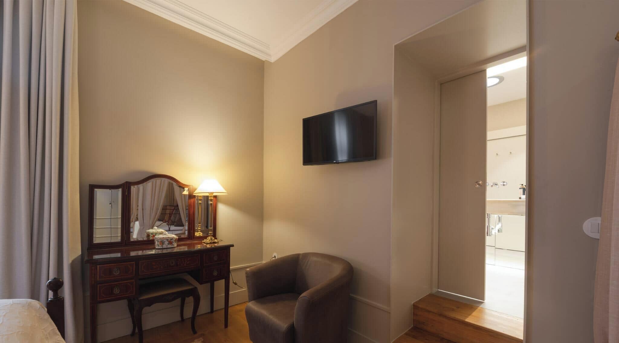 A hotel room with a bed, a desk with a lamp, a chair, a TV mounted on the wall, and a bathroom visible through an open door.