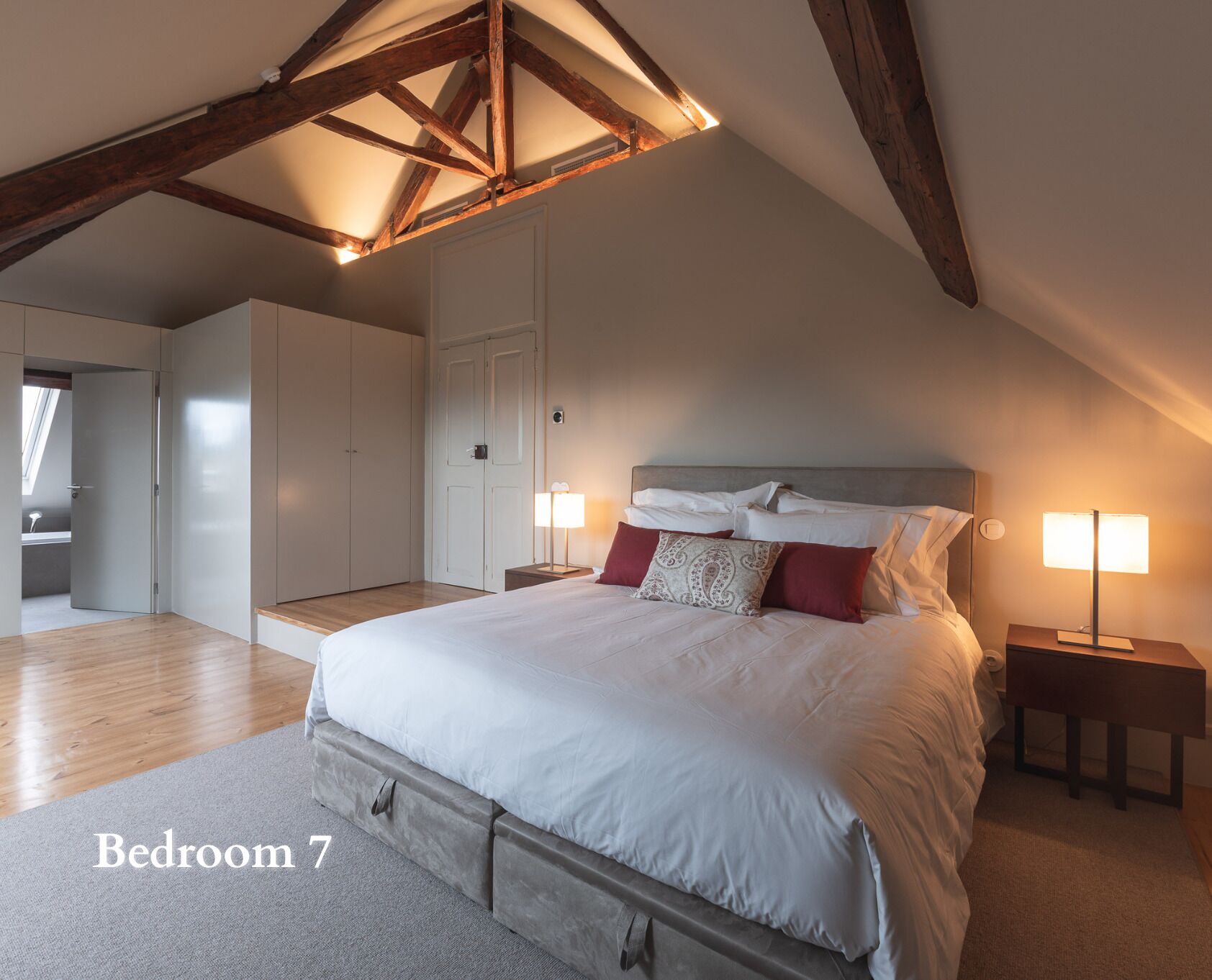 A bedroom with a large bed, wooden beams, a skylight, and a view of another room.