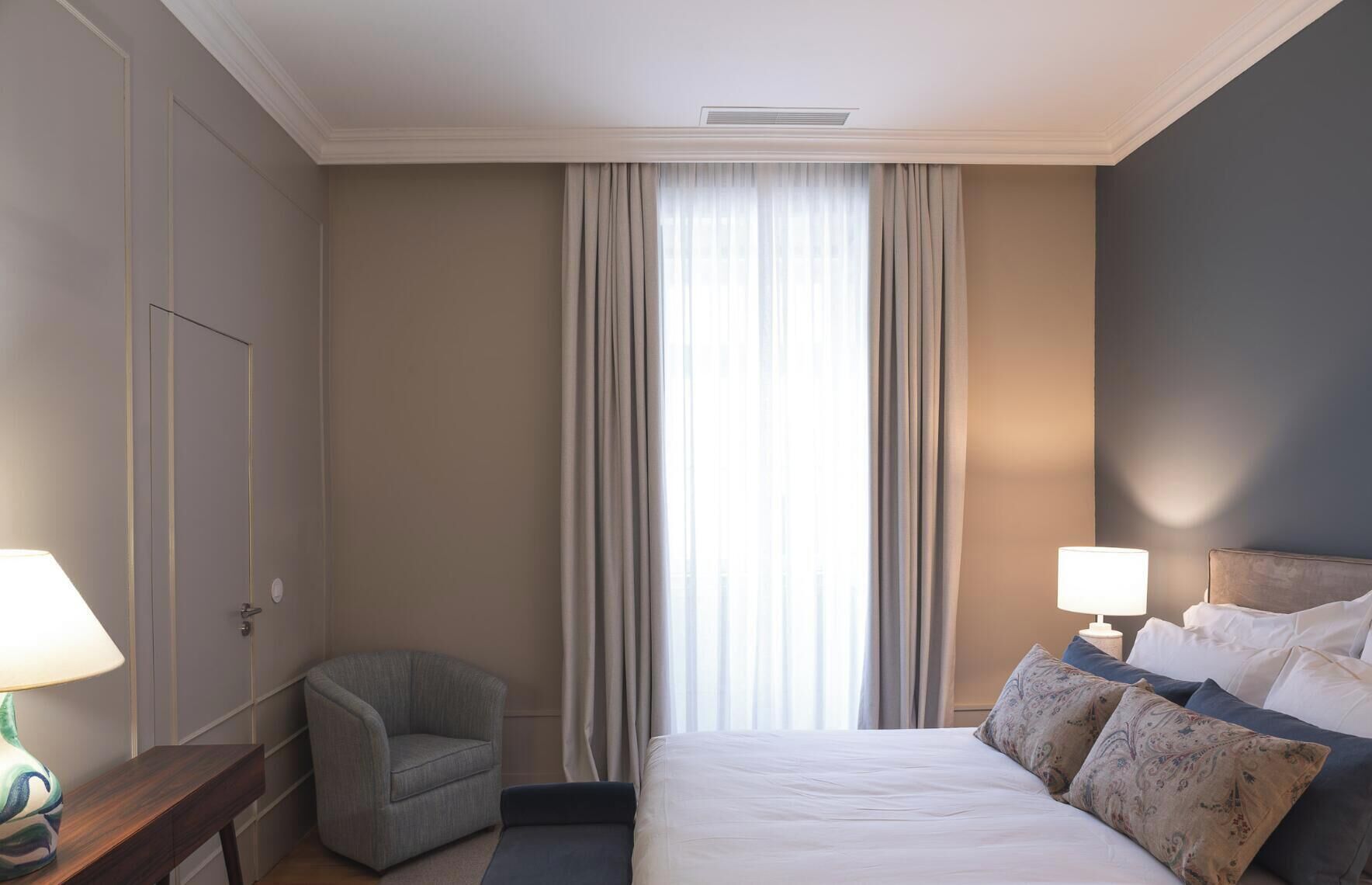 A hotel room with a bed, two bedside lamps, a chair, a nightstand, and a window with curtains.