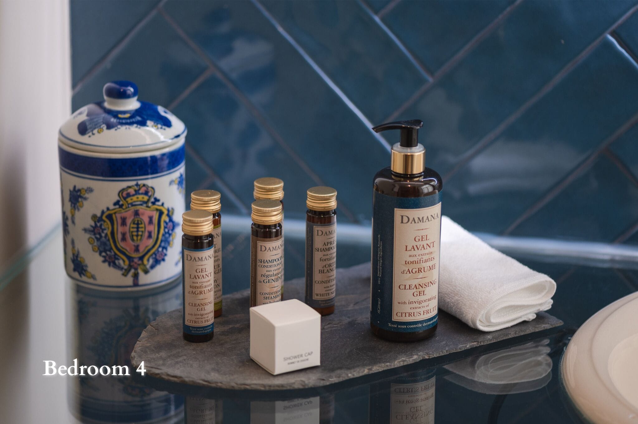 A bathroom with a blue tiled wall, a white ceramic container with a blue lid, and various toiletries including a bottle of shampoo, a bottle of conditioner, and a shower cap.