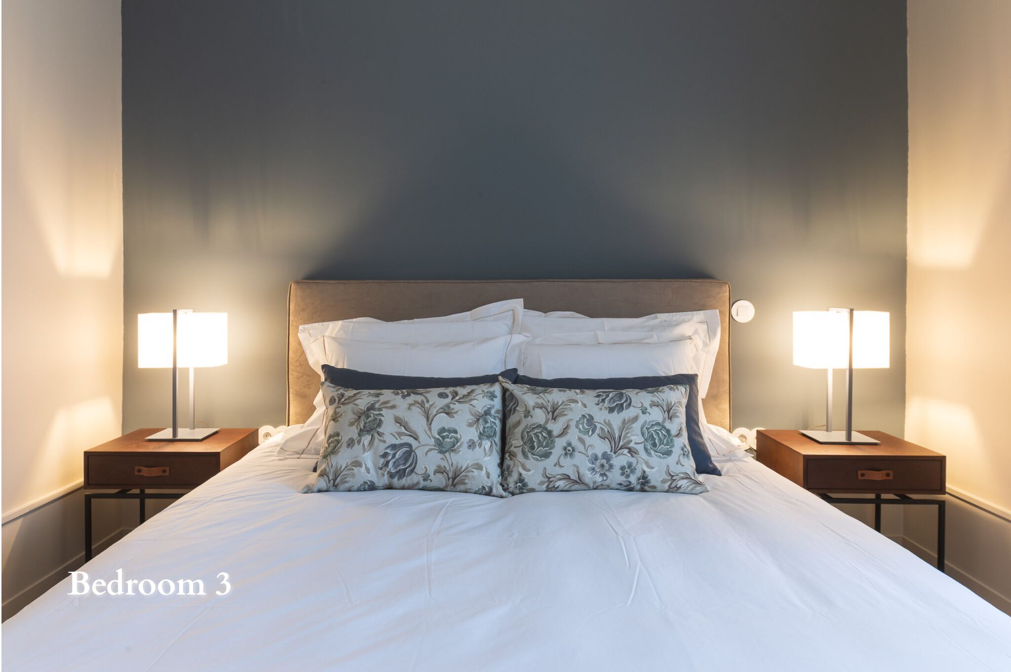 A neatly made bed with floral pillows in a bedroom with two bedside lamps and a nightstand.