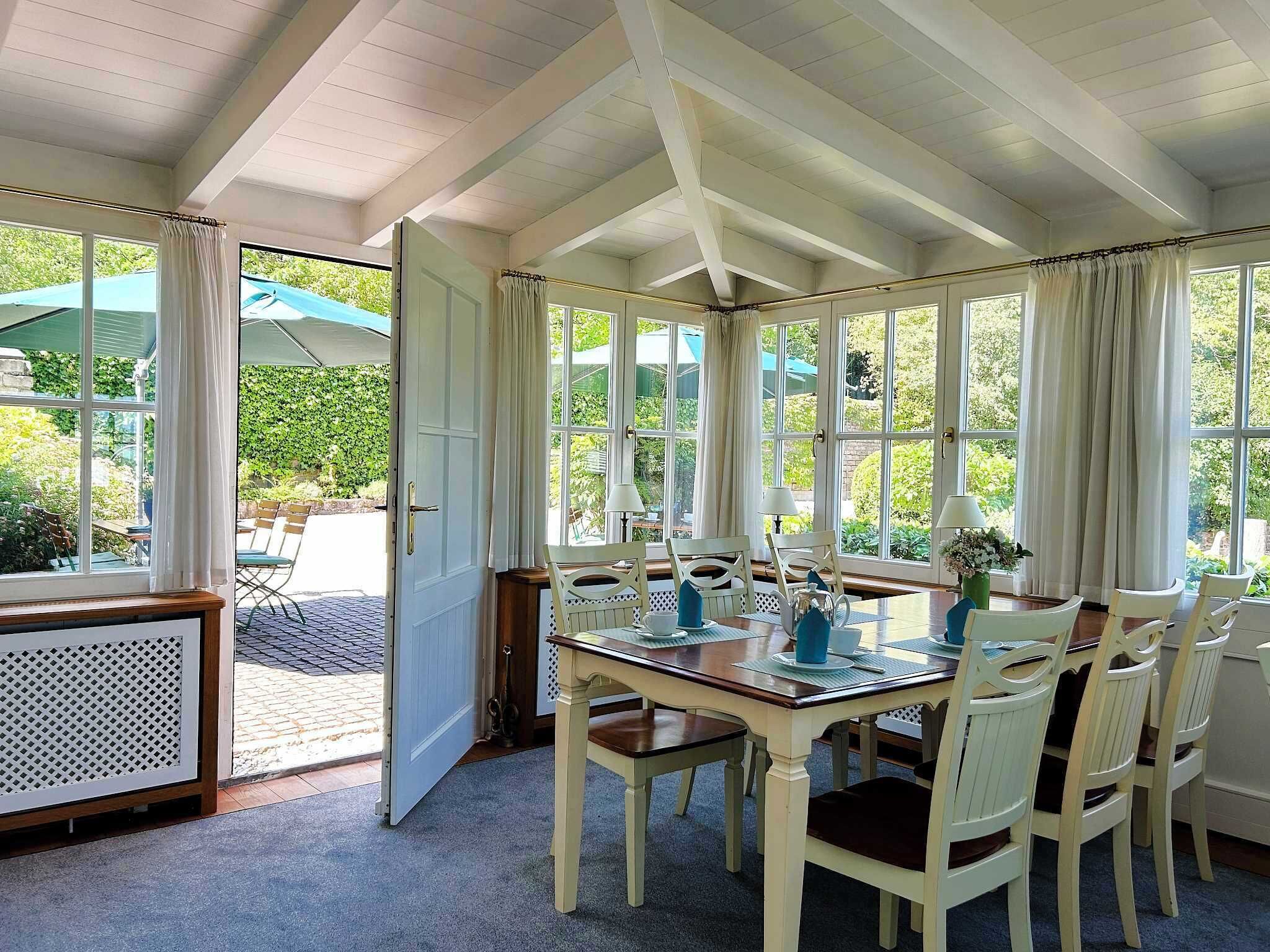 A dining area with a wooden table and white chairs, large windows with curtains, and a view of greenery outside.