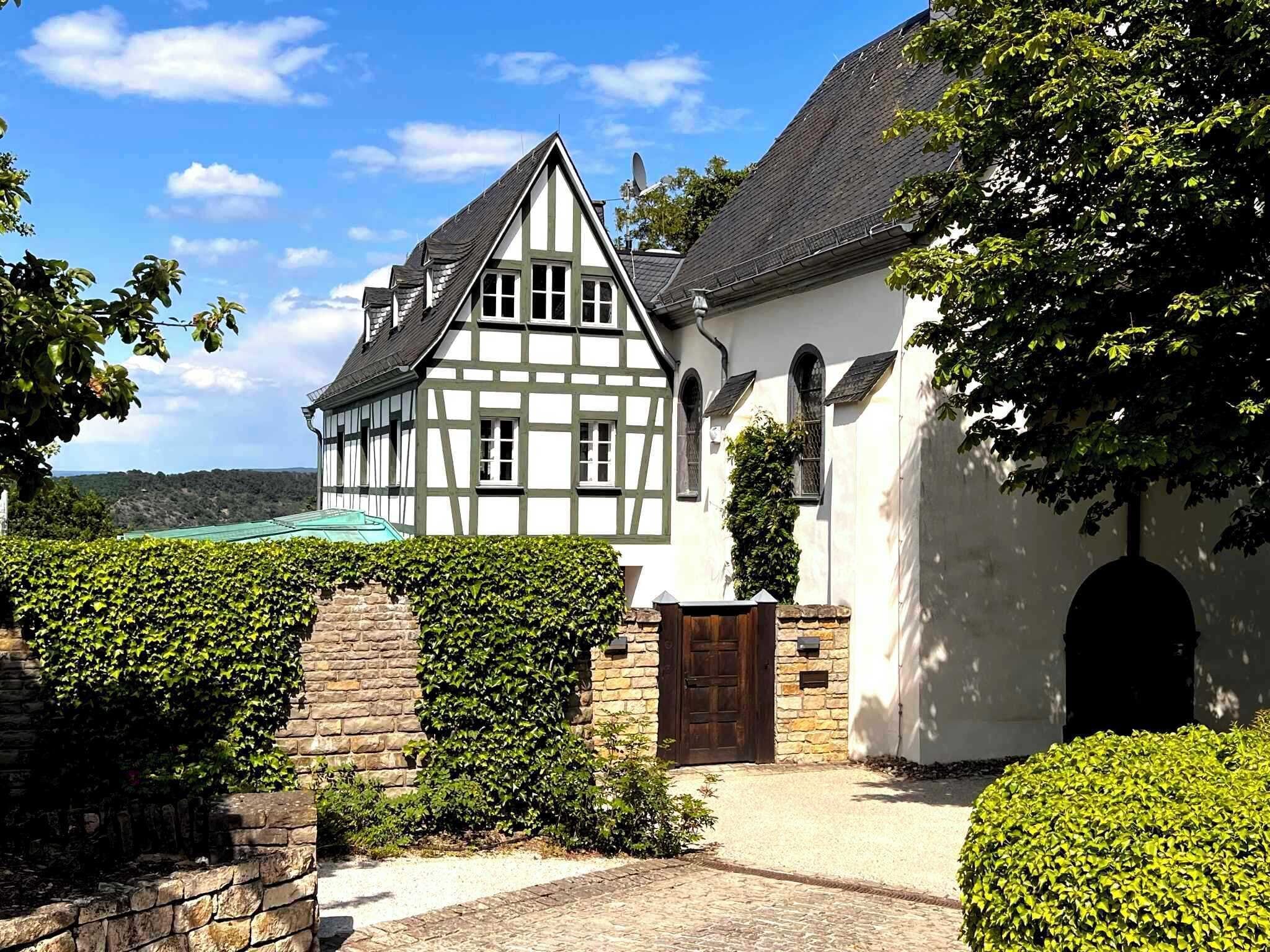 A traditional half-timbered house with a stone wall and a wooden door, surrounded by well-maintained hedges and a cobblestone path.