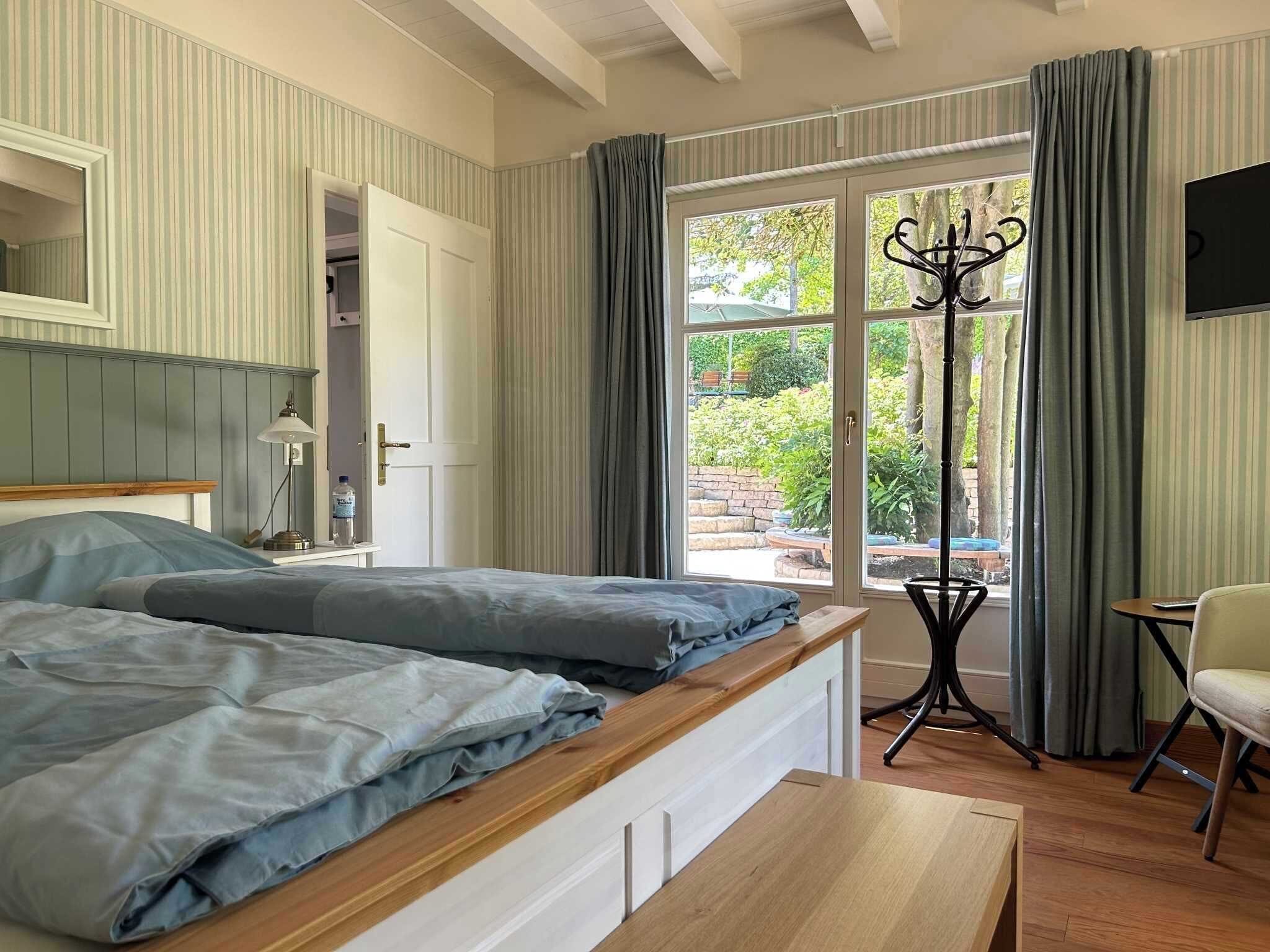 A bedroom with a bed, bedside table, lamp, and a view of the outdoors through a window.