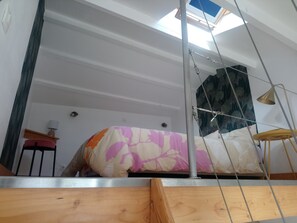 A high-ceiling room with a skylight, a bed with patterned bedding, a wooden chair, and a small table.