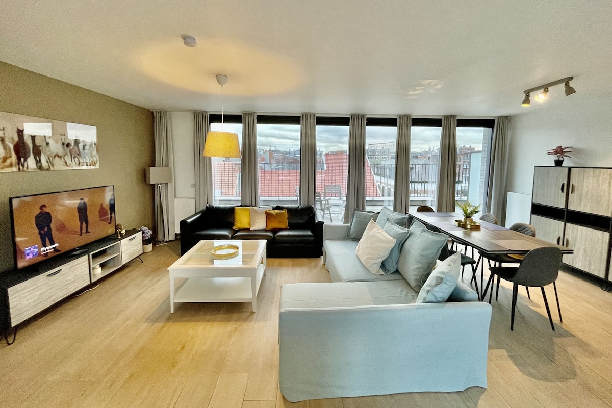 Een moderne woonkamer met een grote televisie, banken, een eettafel en uitzicht op gebouwen door grote ramen.