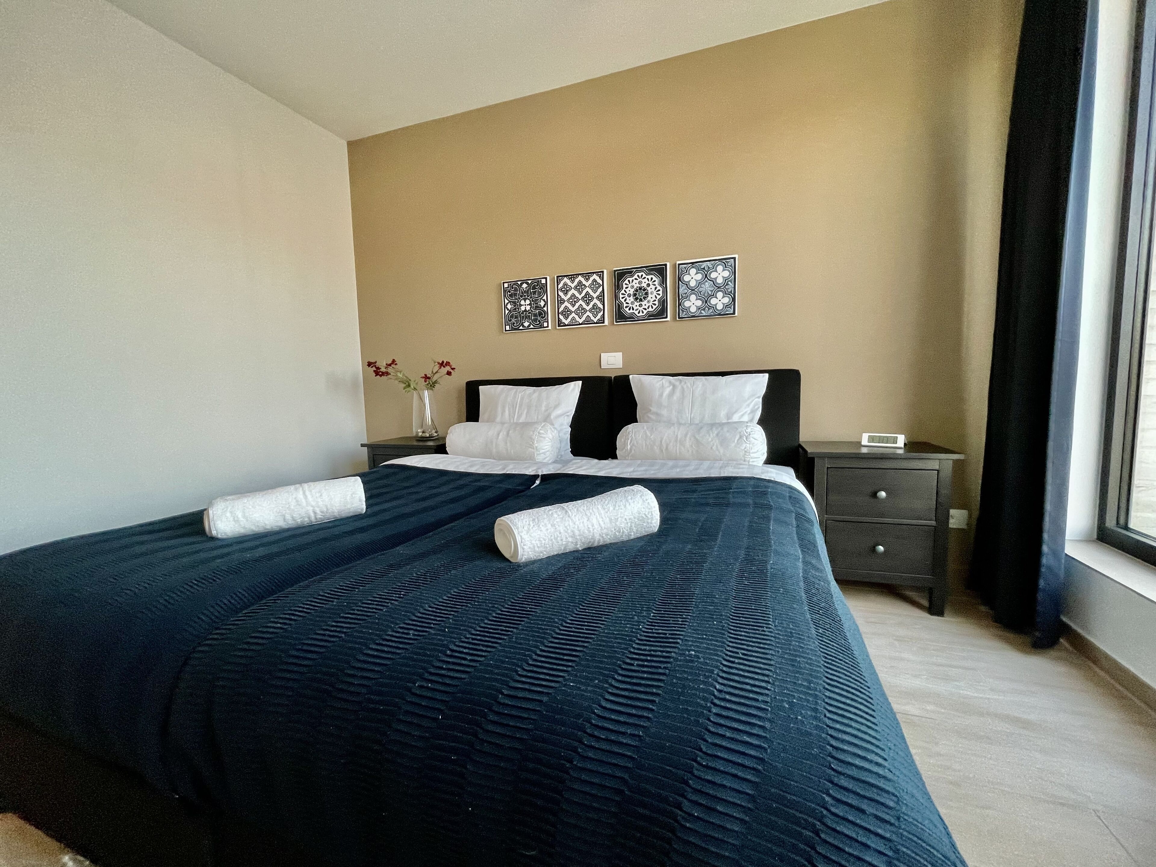 Una cama bien hecha con una colcha azul y almohadas blancas, una mesita de noche con un florero y una pared con tres platos decorativos.