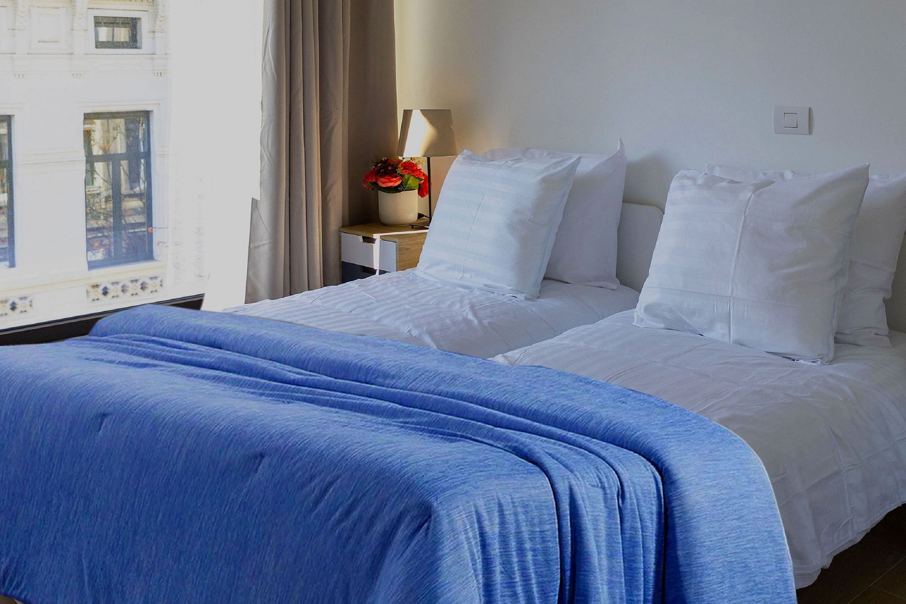 Habitació d'hotel amb un llit, una tauleta de nit amb una làmpada i flors, i una finestra amb cortines.