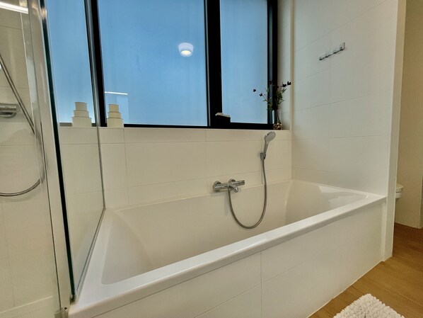 A modern bathroom with a glass shower enclosure, a white bathtub, and a window with a view of the sky.