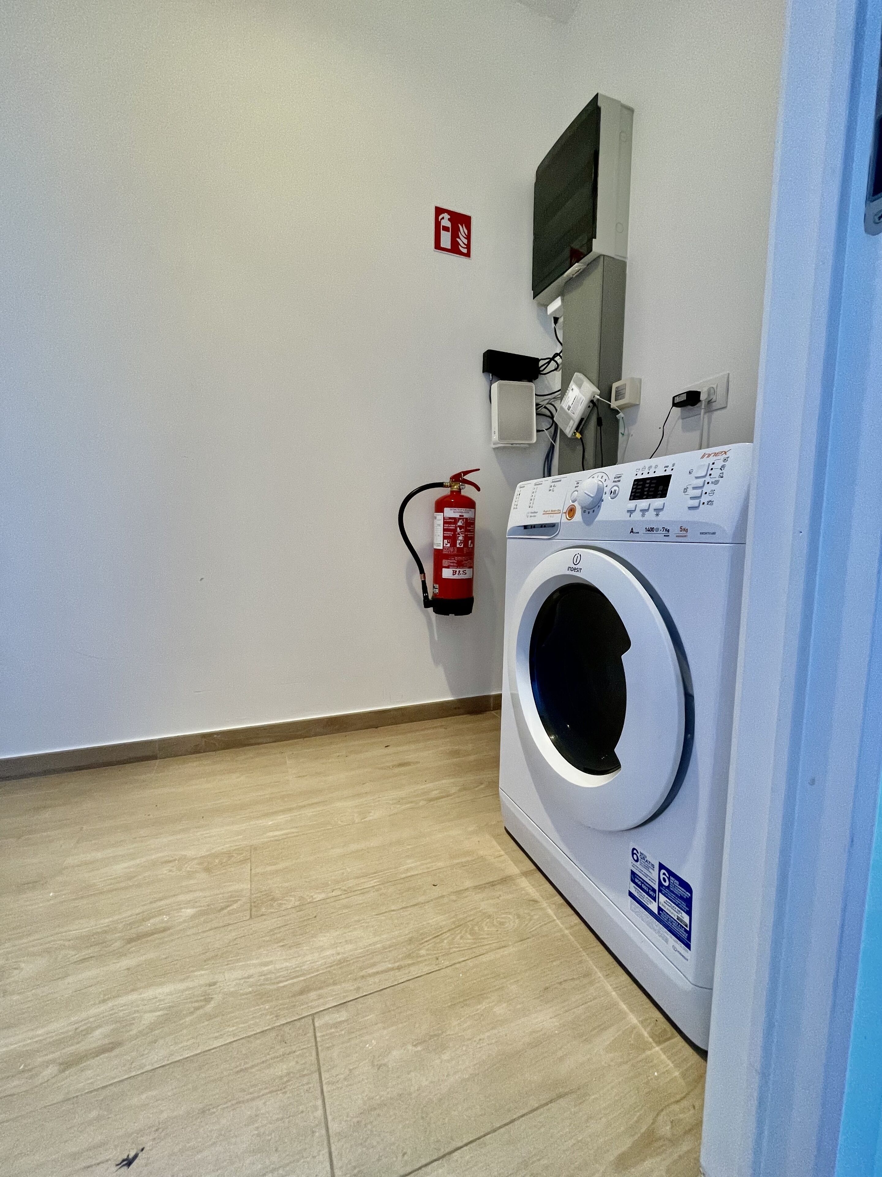 A laundry room with a front-loading washing machine, a fire extinguisher mounted on the wall, and a power outlet.