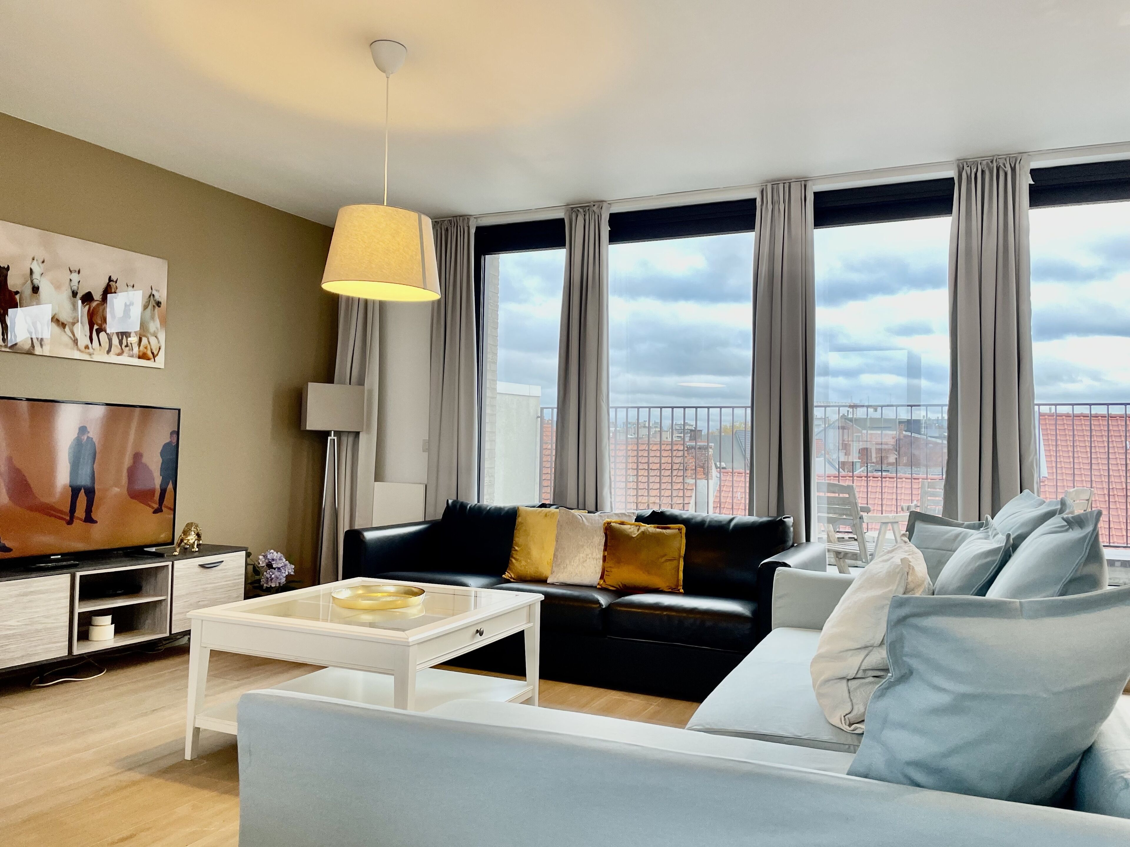 A modern living room with a black leather sofa, a white coffee table, a flat-screen TV, and large windows with curtains.