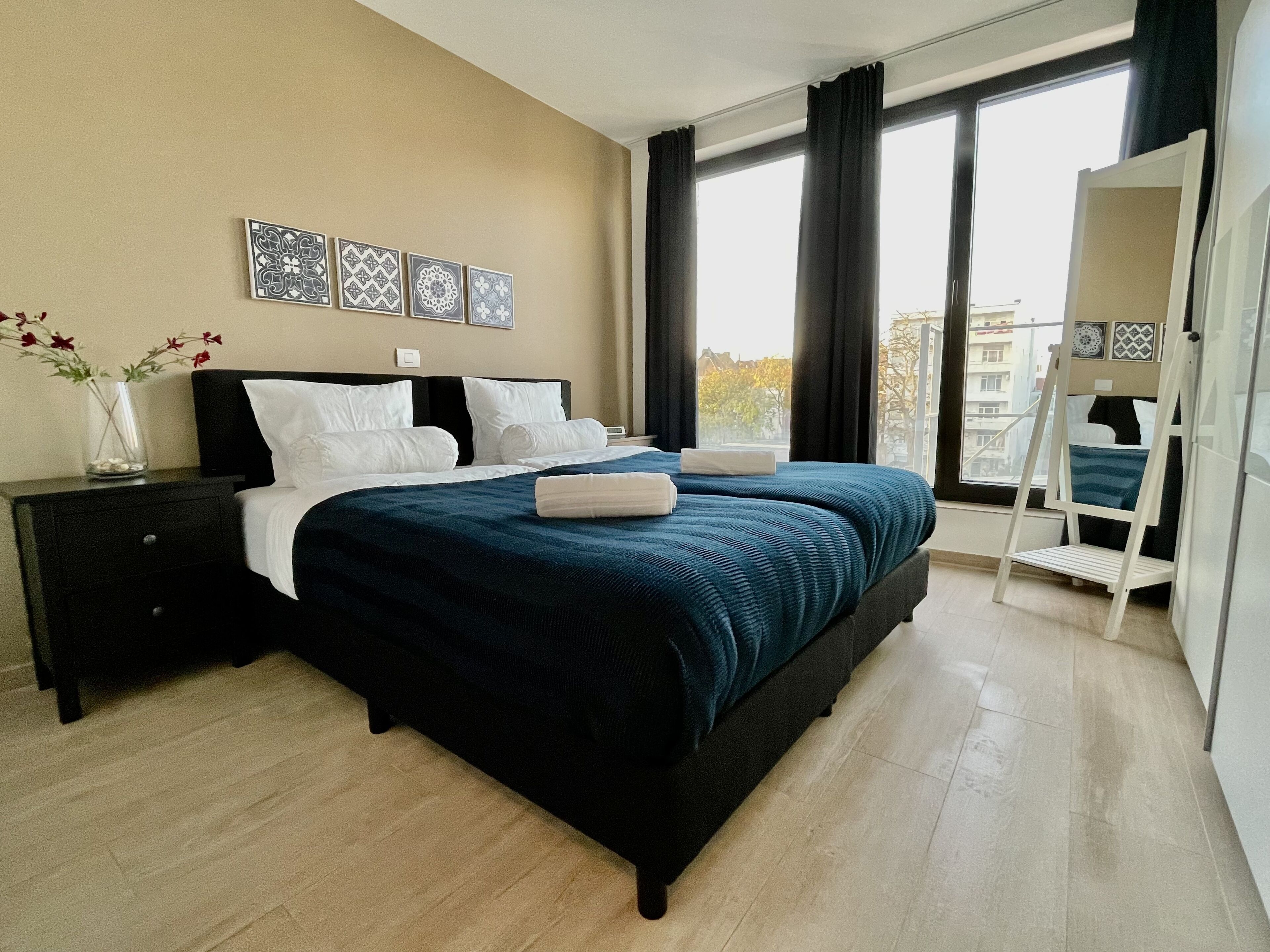 Une chambre à coucher moderne avec un grand lit, une table de chevet, une commode et une vue sur la ville grâce à de grandes fenêtres.
