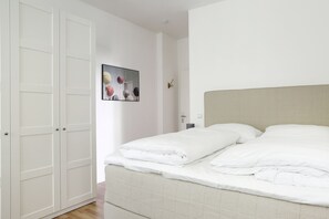 Cama bem arrumada com lençóis brancos em quarto com armário branco fechado e quadro fixado na parede.