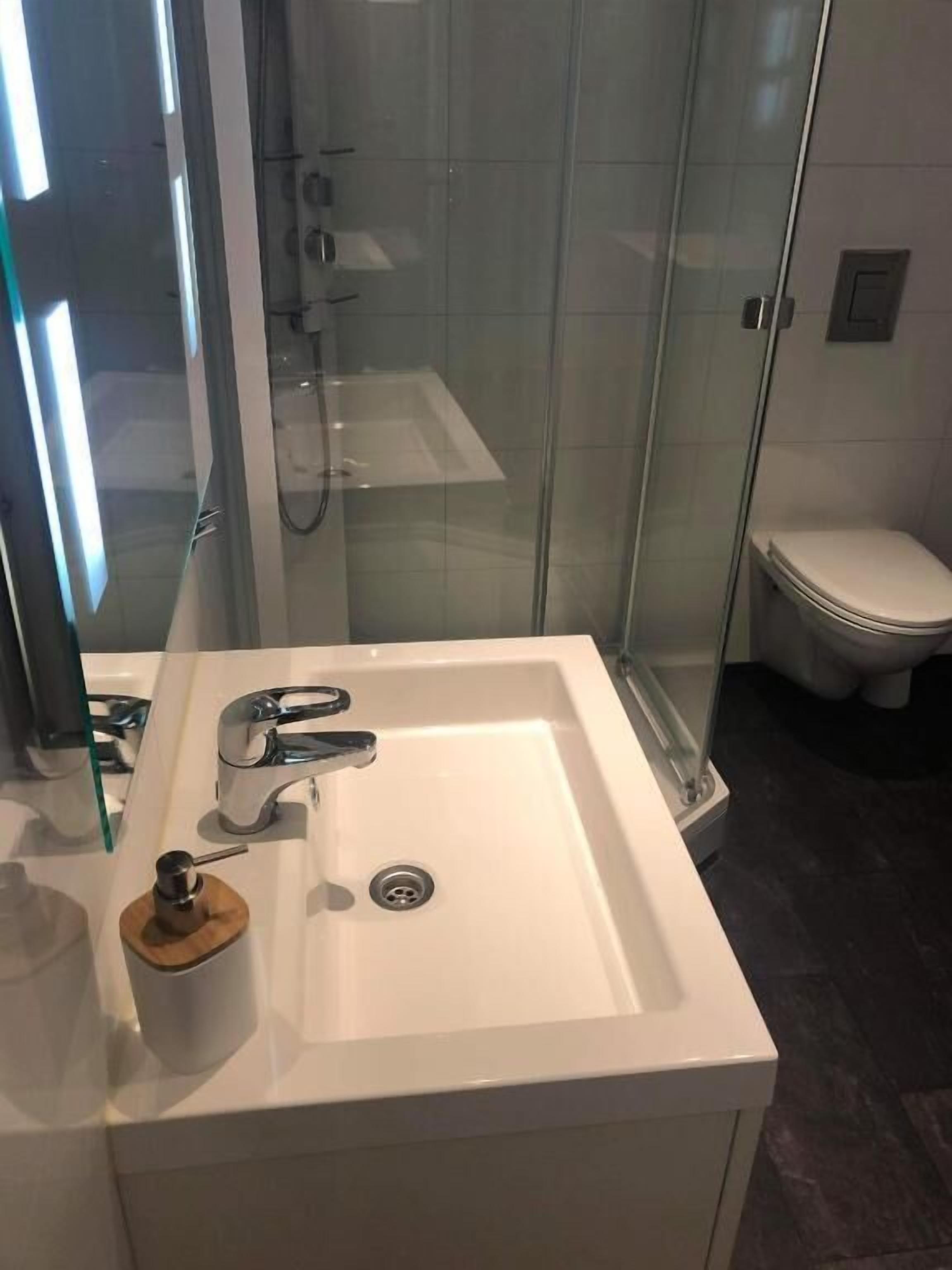 Un baño moderno con ducha envuelta en vidrio, un lavamanos blanco y un dispensador de jabón de madera, y un inodoro.