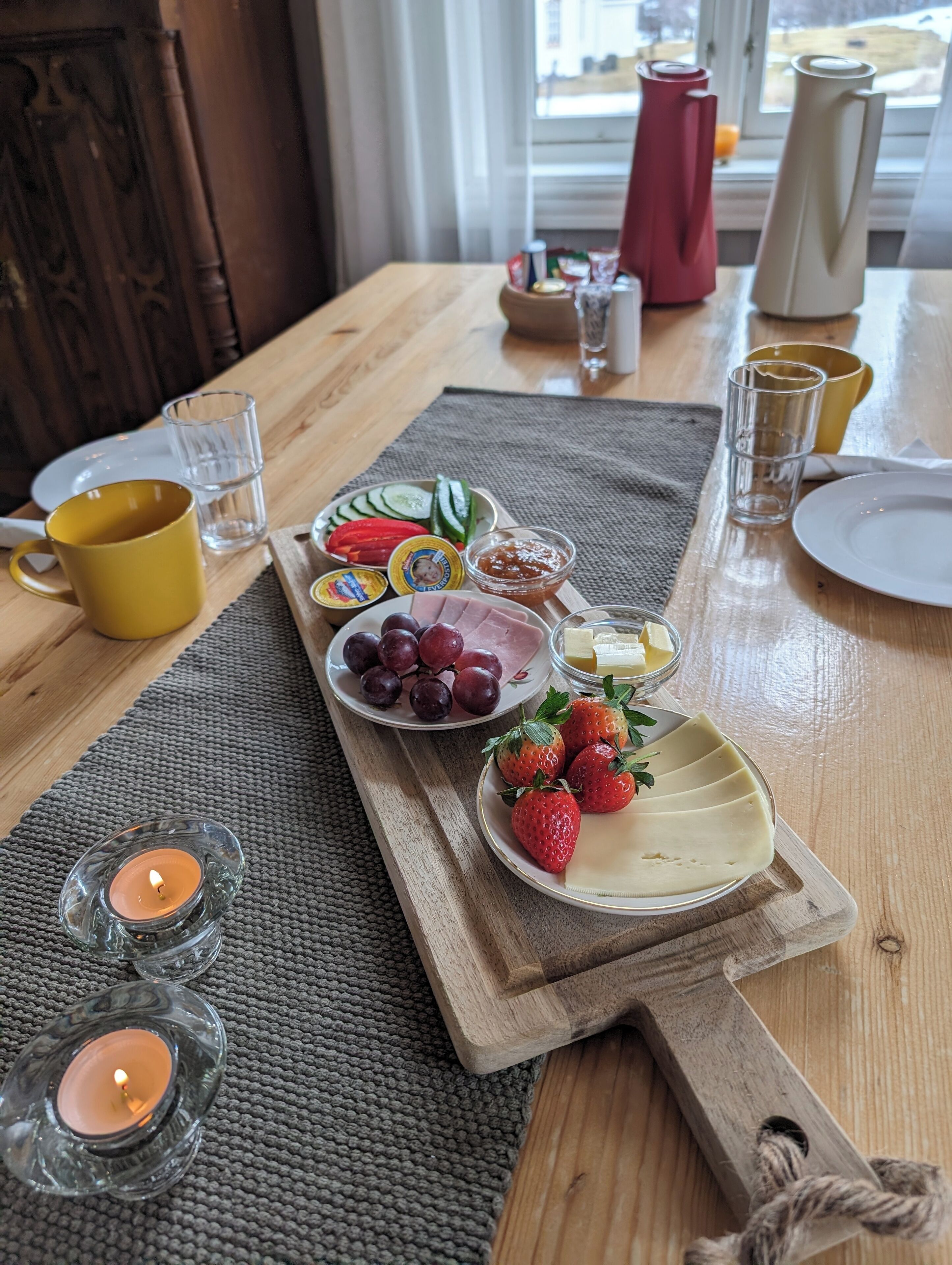 Una mesa de madera con una variedad de alimentos y bebidas, incluyendo platos de uvas, fresas y verduras en rodajas, junto con una taza amarilla y vasos.