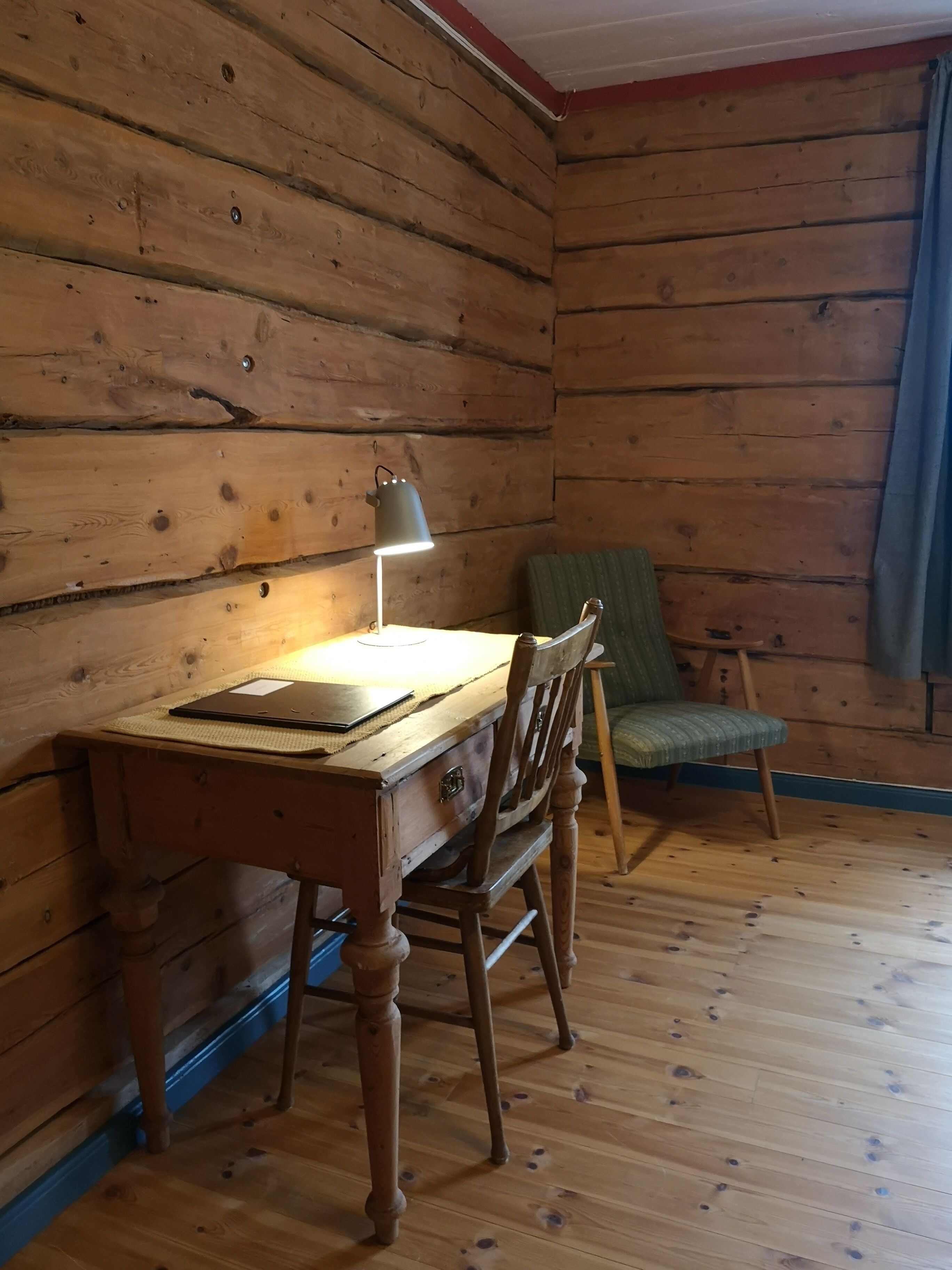 Interno di una baita di legno con una scrivania, una sedia, una lampada e una finestra con tende.