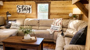 Ein gemütliches Wohnzimmer mit einer Holzwand, einer Sofagarnitur, einem Couchtisch mit Vase und Kerze sowie einem Schild mit der Aufschrift 'Stay'.