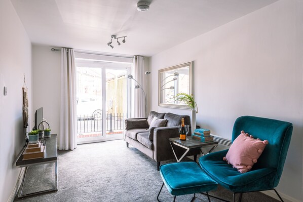 A modern living room with a grey sofa, a teal armchair, a glass coffee table, and a small side table with a bottle and glasses.