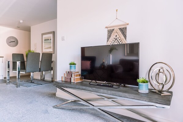 A modern living room with a flat-screen TV, two grey chairs, a coffee table, and decorative items including a plant and a geometric wall hanging.