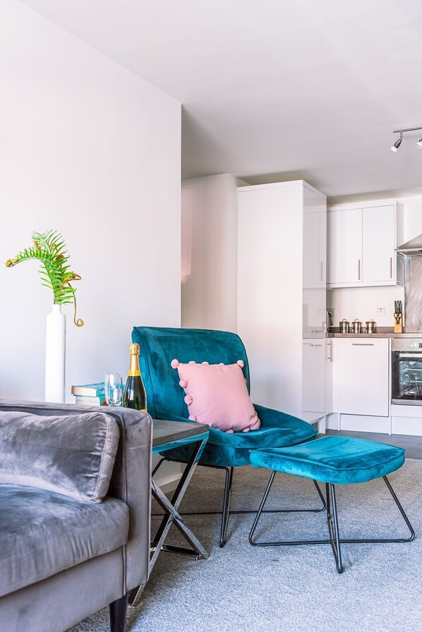 A modern living room with a teal armchair, a grey sofa, and a small table with a bottle and glass. A white cabinet and a kitchen area are visible in the background.