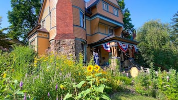 Uma casa de dois andares com um canto curvo, chaminé de tijolos e varanda frontal com bandeiras americanas.