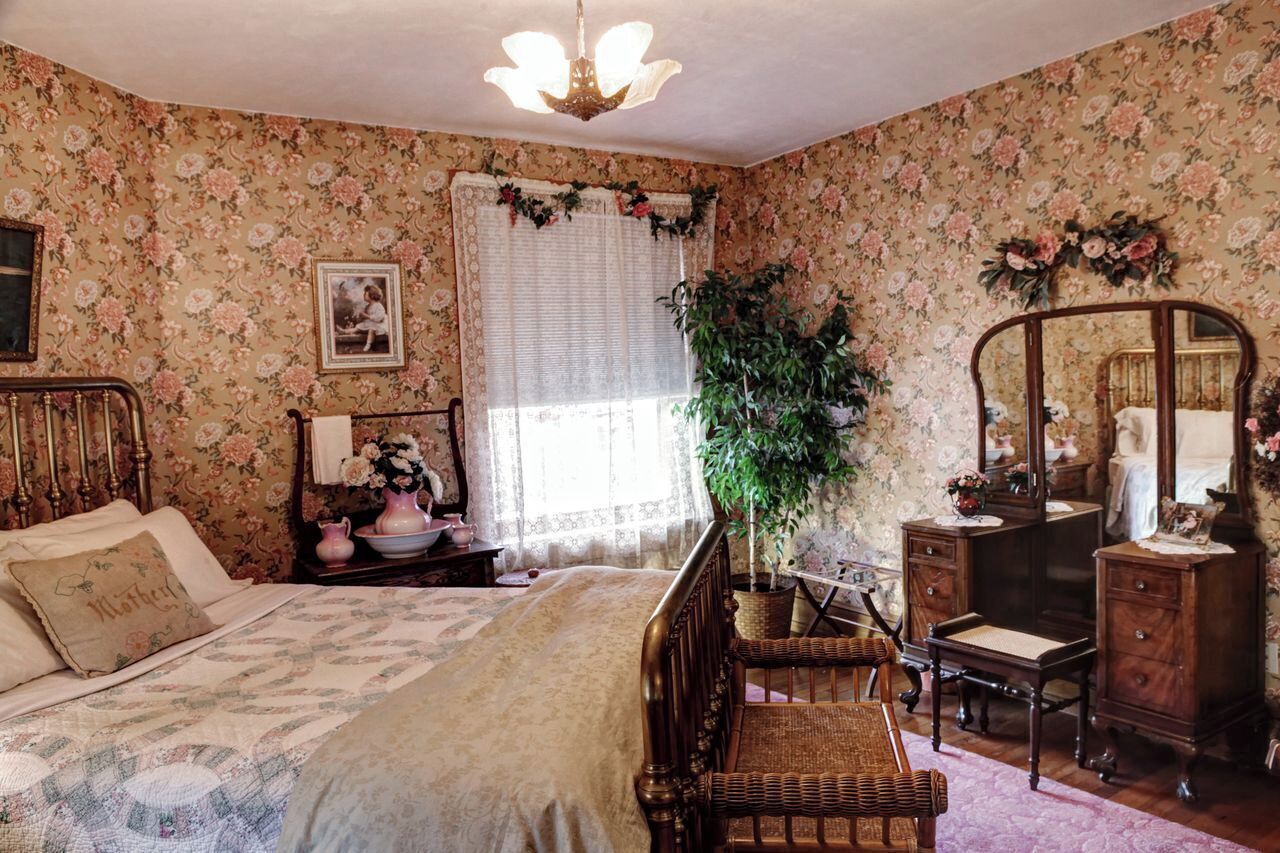 Ein Schlafzimmer mit floraler Tapete, einem Bett mit Holz Kopfteil, einem Schminktisch, einem Spiegel und einem Blumentopf.