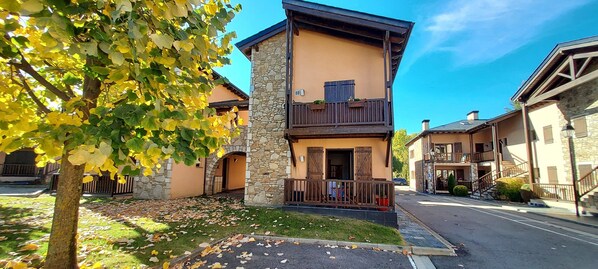A two-story building with stone and wooden exterior, a balcony, and a tree with yellow leaves in the foreground.