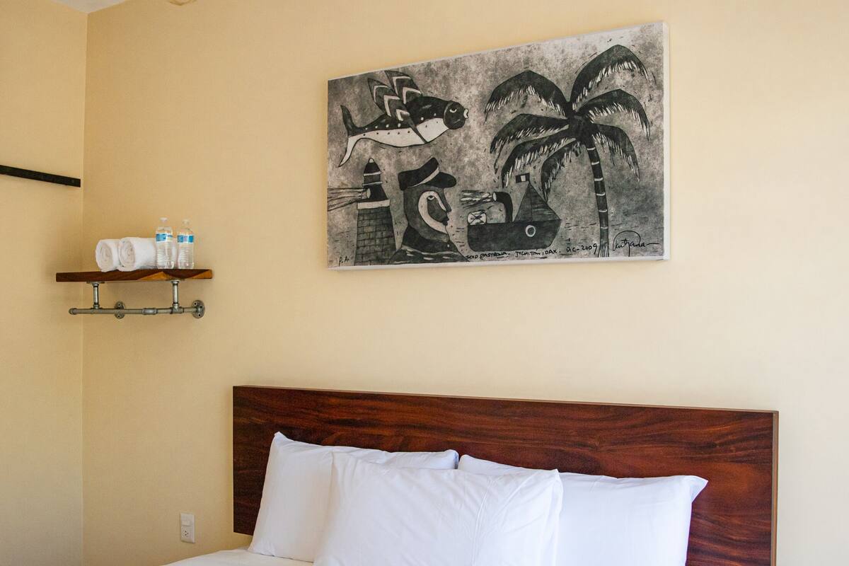 A neatly made bed with white linens, a wooden headboard, and a striped throw blanket. There is a wall-mounted shelf with towels and toiletries. A painting of palm trees and a boat hangs above the bed.
