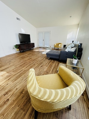 A modern living room with a wooden floor, a sofa, a yellow armchair, a TV, and a glass coffee table.