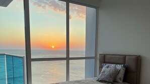 Een slaapkamer met een groot raam met uitzicht op zee, een bed met beddengoed met een patroon en een balkon met een glazen balustrade.