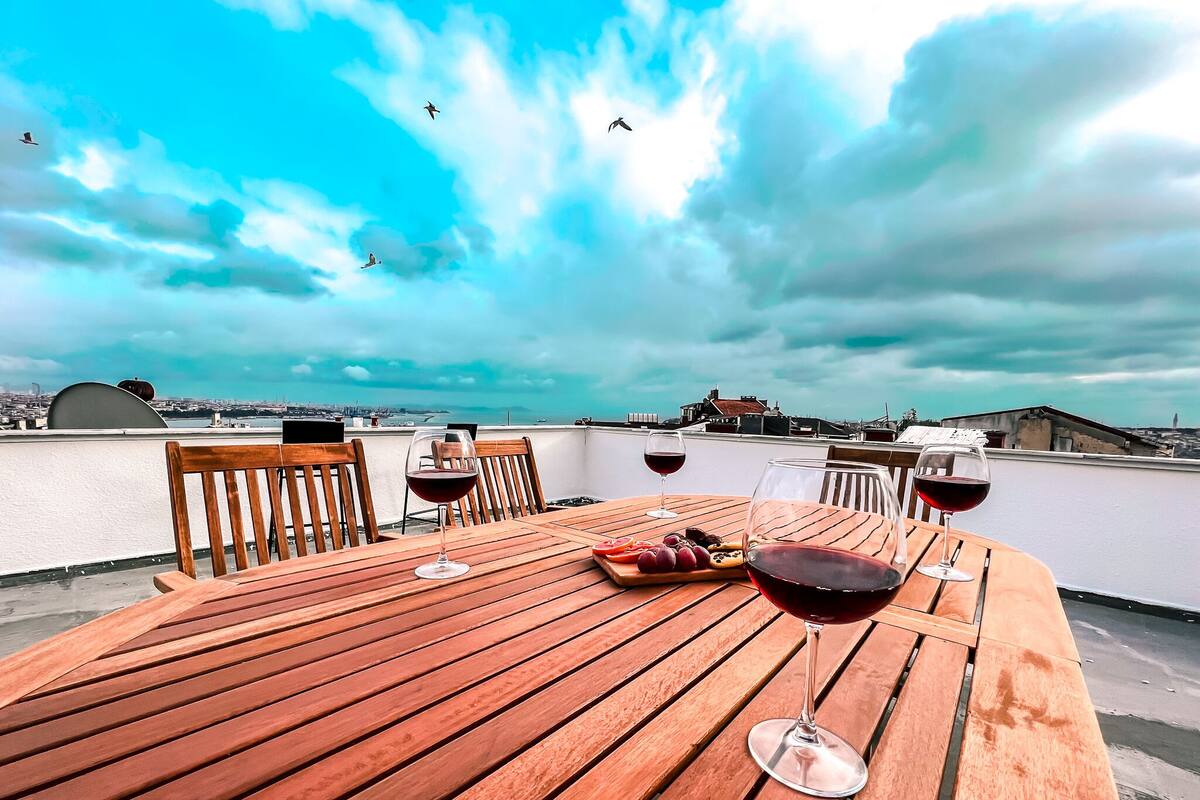 A wooden table with two wine glasses and a plate of grapes on a rooftop terrace with a city view.