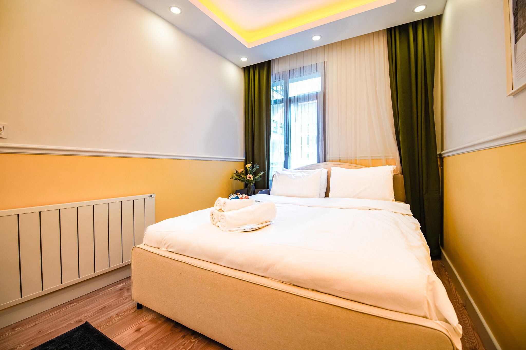 A neatly made bed with white linens, a radiator, and a black rug on a wooden floor.