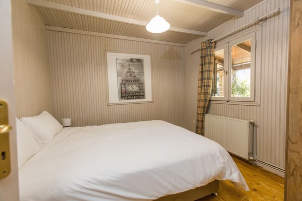Una cama bien hecha en una habitación con un radiador, una ventana con cortina escocesa y un cartel del London Eye en la pared.