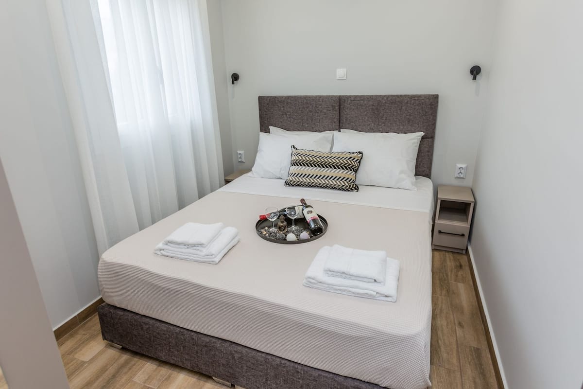 Un lit bien fait, avec une tête de lit grise, des oreillers blancs et un coussin décoratif. On peut voir des serviettes pliées sur le lit et un plateau avec une bouteille et des verres.
