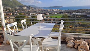 Une terrasse avec une table et des chaises blanches, une lampe, et une vue panoramique sur une ville côtière et les montagnes.