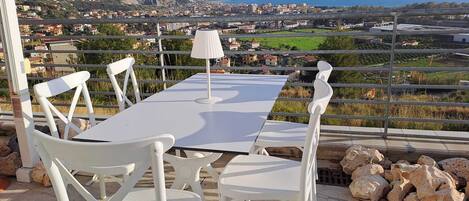 A terrace with a white table and chairs, a lamp, and a scenic view of a coastal town and mountains.