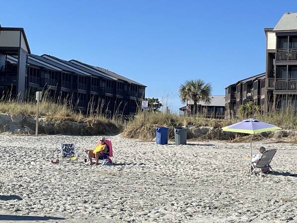 A beach with a chair under a beach umbrella, a person reading, and a building in the background.