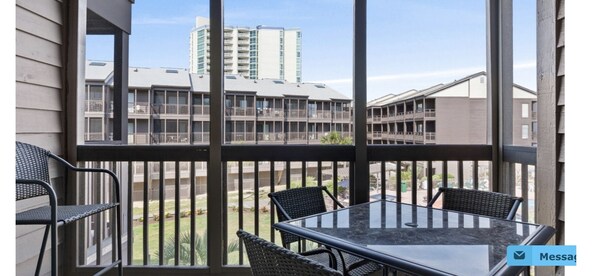 A balcony with a glass table and chairs, overlooking a courtyard with buildings in the background. - Amazing Ocean and Pool Views!  Unit 211 - BEAUTIFUL POOL DECK REMODEL COMPLETE! (North Myrtle Beach)