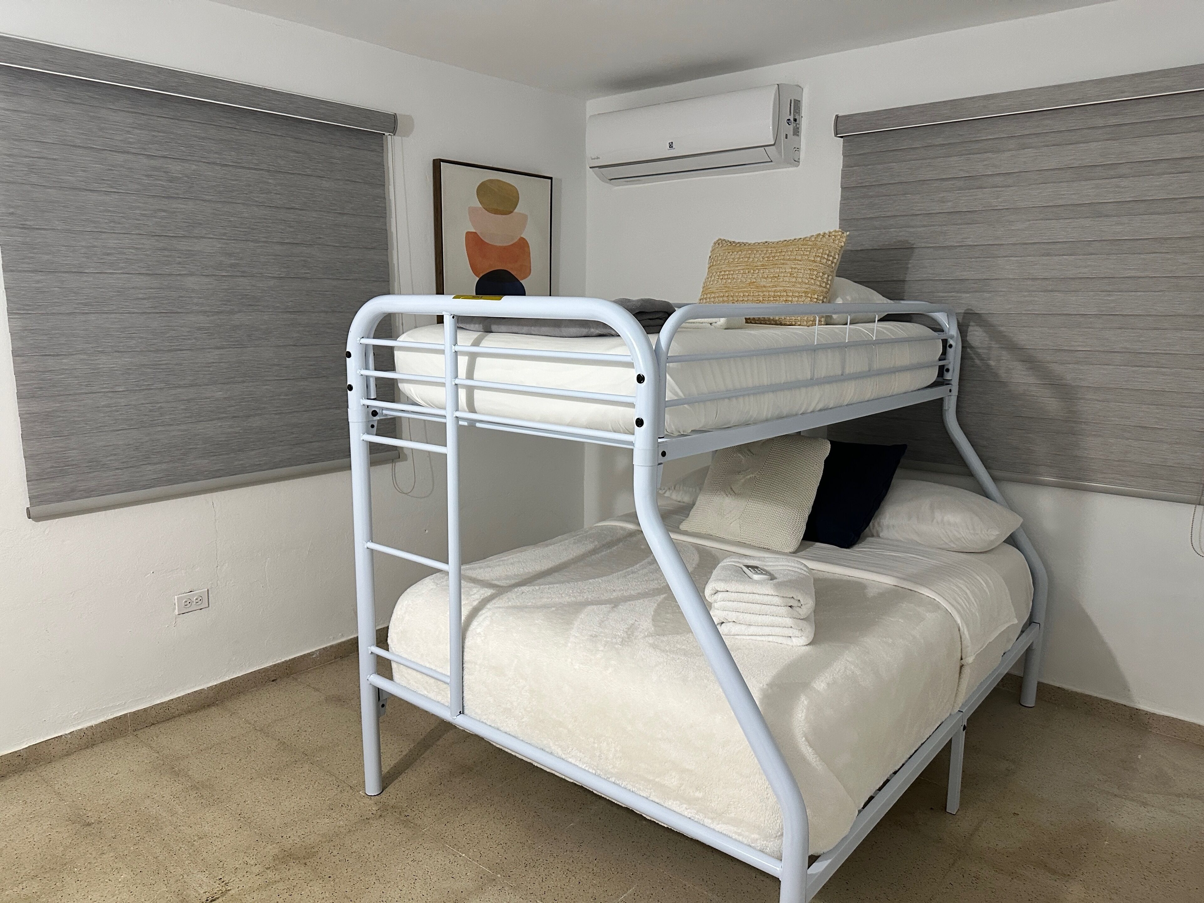 A bunk bed with a metal frame, white bedding, and a single pillow. There is a window with blinds, an air conditioning unit on the wall, and a framed picture above the bed.