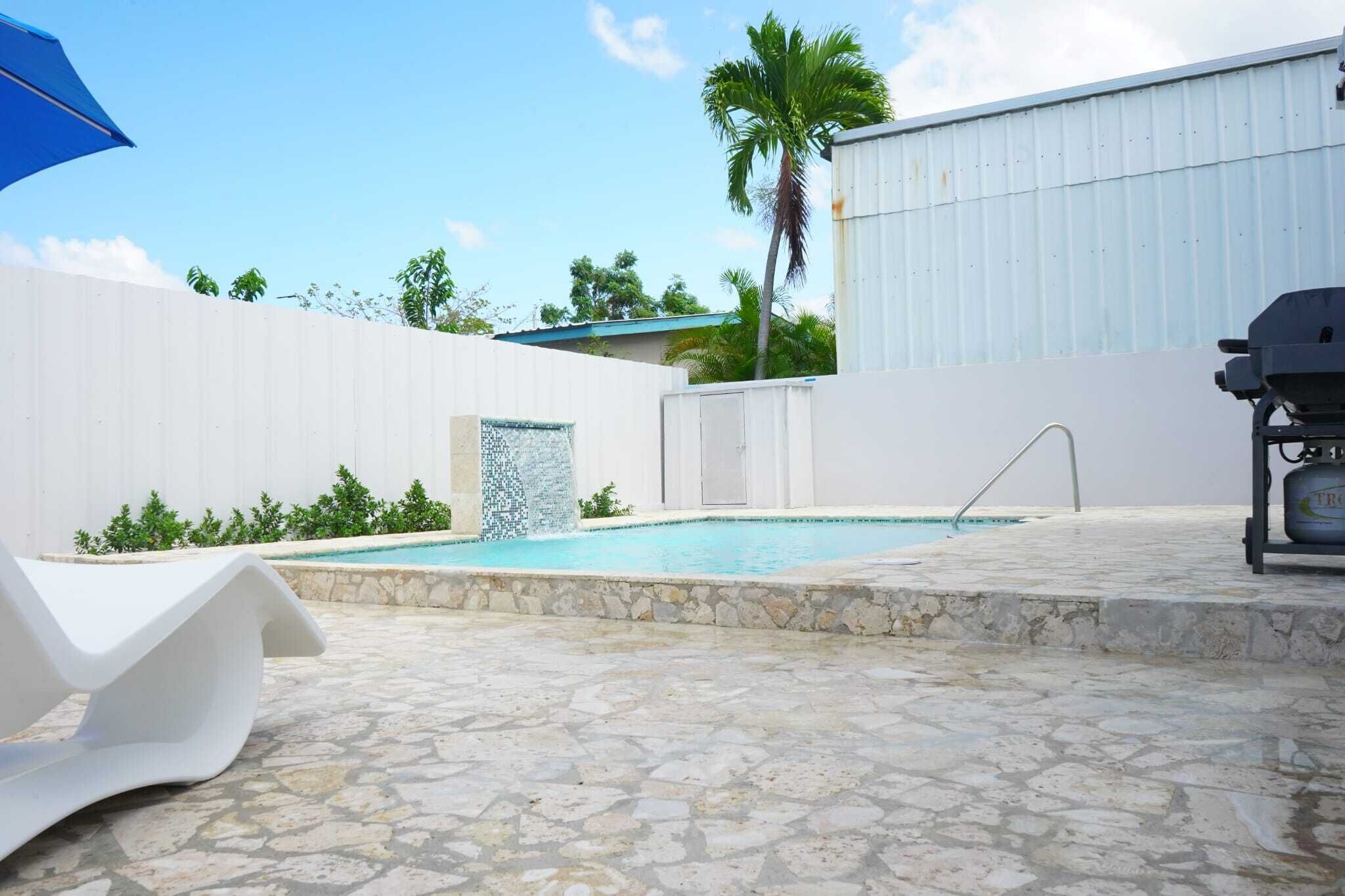 Una piscina con deck de piedra, pared blanca y una parrilla.