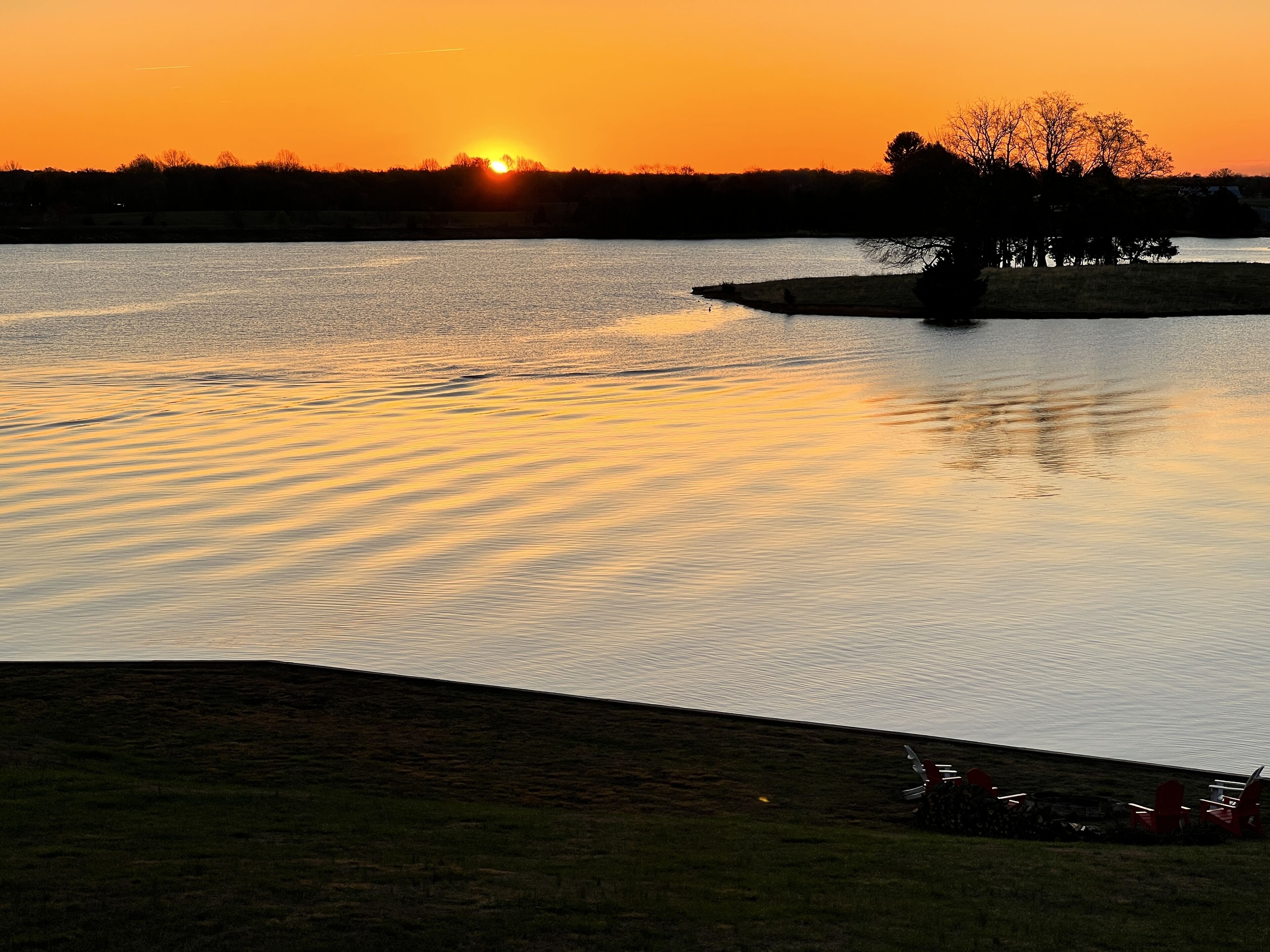 A sunset over a lake with a grassy area and a few chairs.